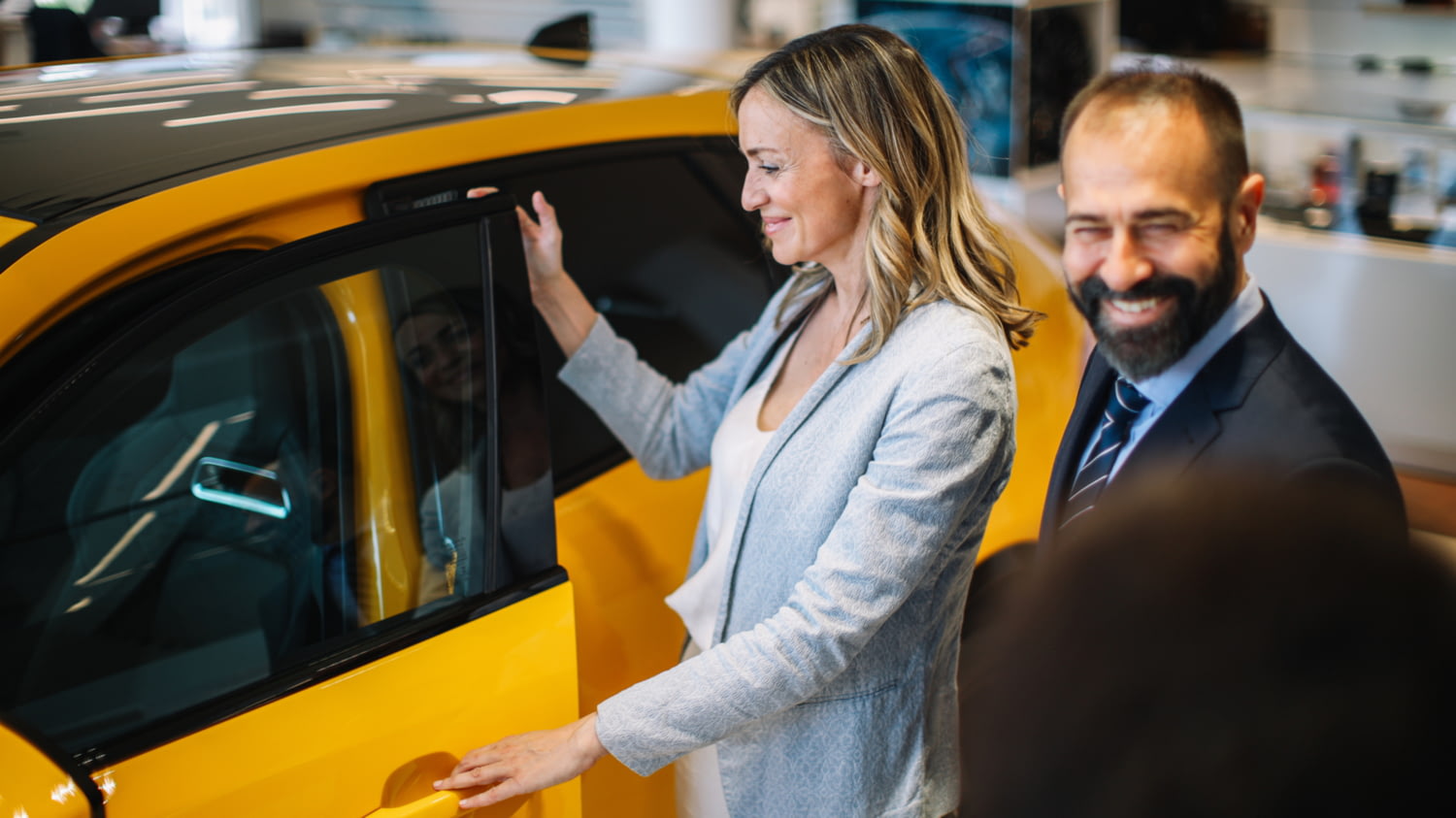 Eine blonde Frau öffnet in einem Autohaus lächelnd die Tür eines gelben SUVs