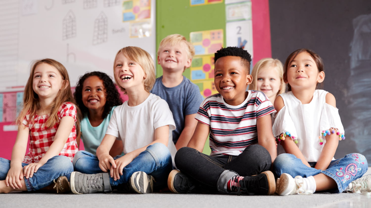 Sieben Kinder sitzen lächelnd auf dem Boden in einem Klassenzimmer