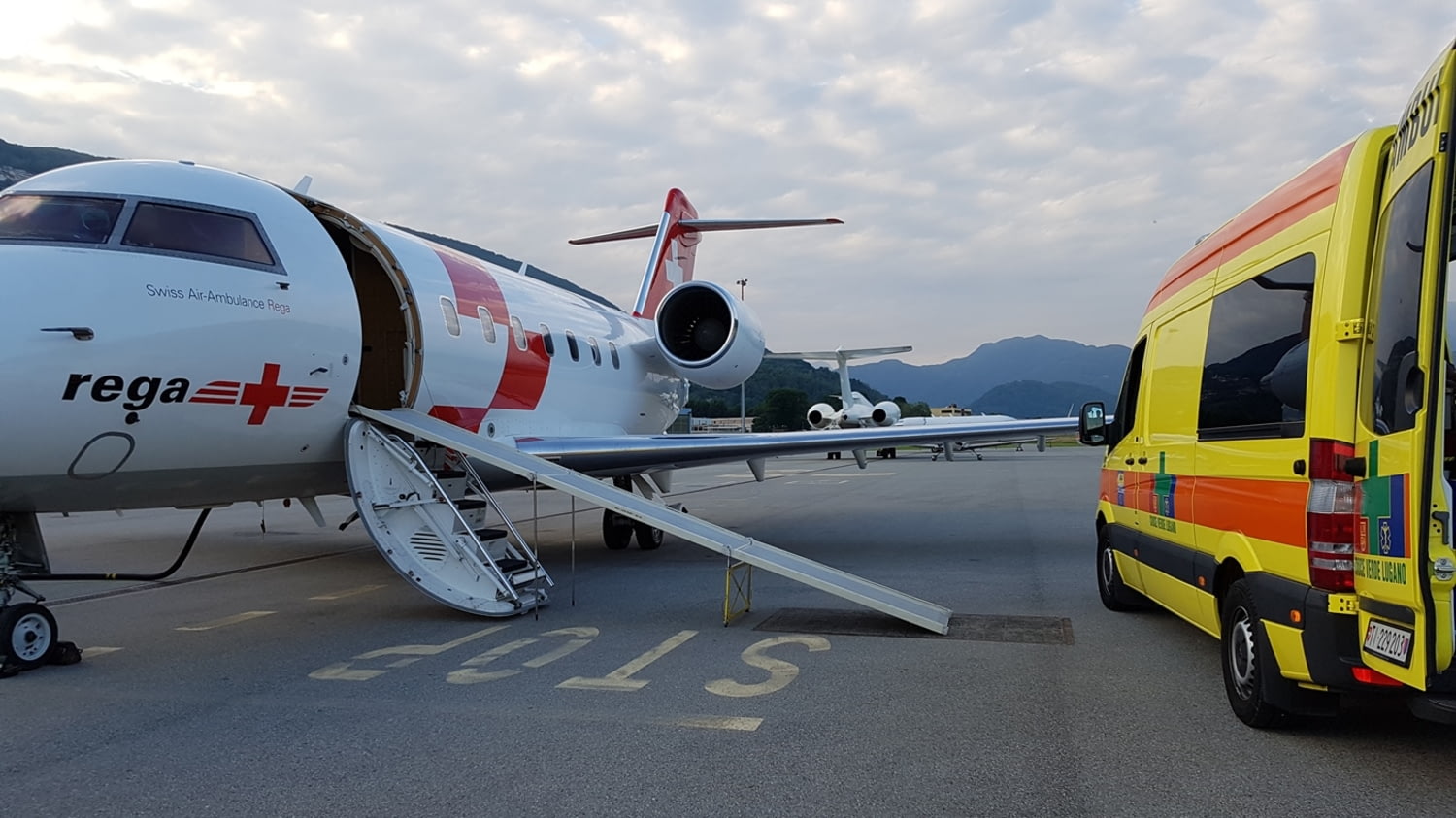 Schweizer Ambulanzflugzeug und ein Rettungswagen aus einem Rollfeld vor Alpenpanorama.