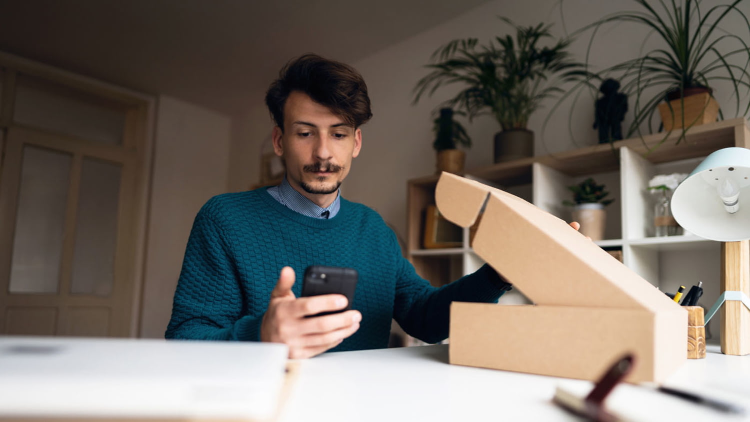 Junger Mann mit geöffnetem Paket vor sich und Smartphone in der Hand