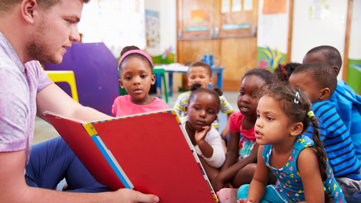 Junger Mann in der Entwicklungshilfe liest einer Gruppe dunkelhäutiger Kinder etwas vor